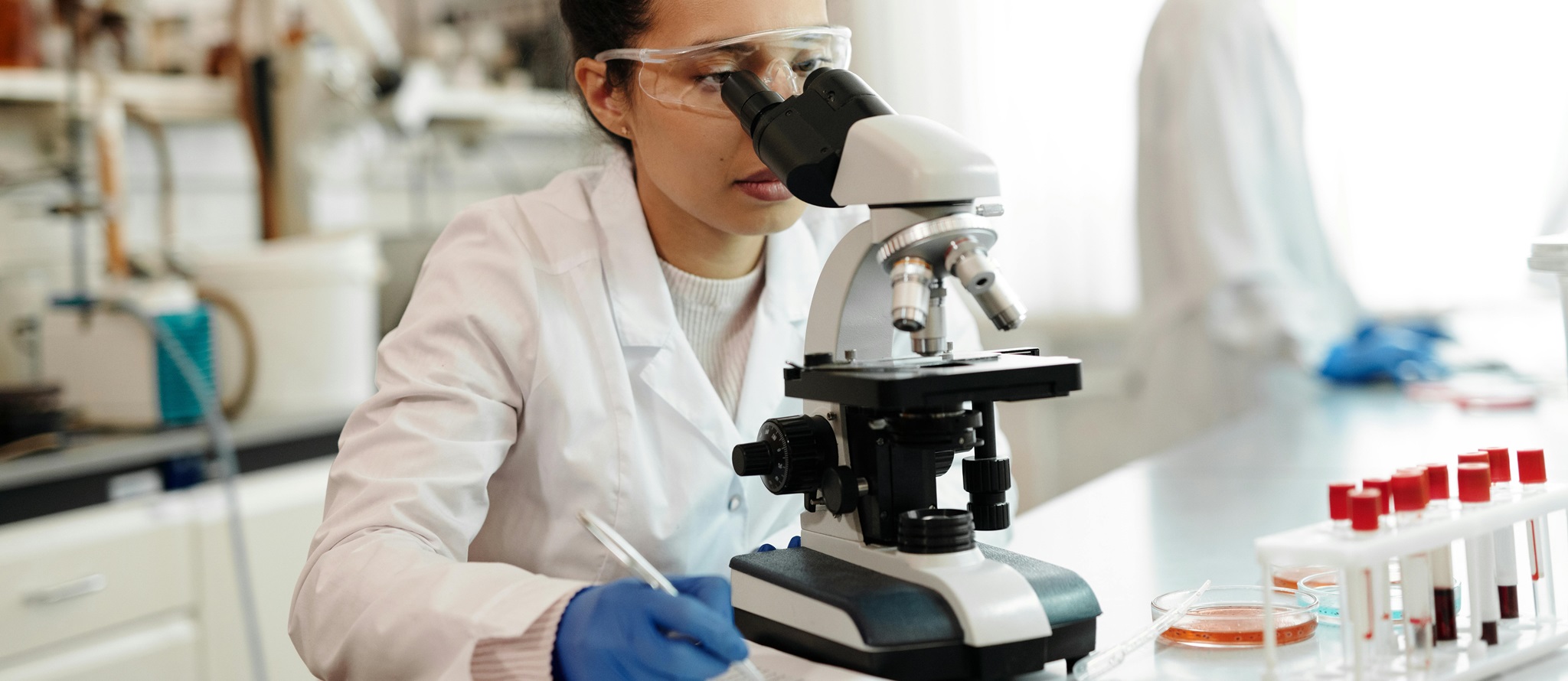 A photo of a female researcher looking through a microscope in a lab, while writing down what she sees. In the back there is another researcher visible wearing a white lab coat.
